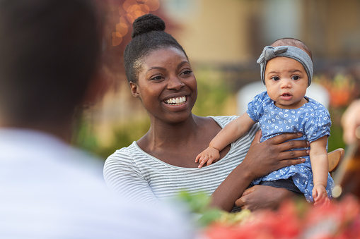 A young African American mom holds her adorable young daughter in her lap at an outdoor dining table with a group of friends.